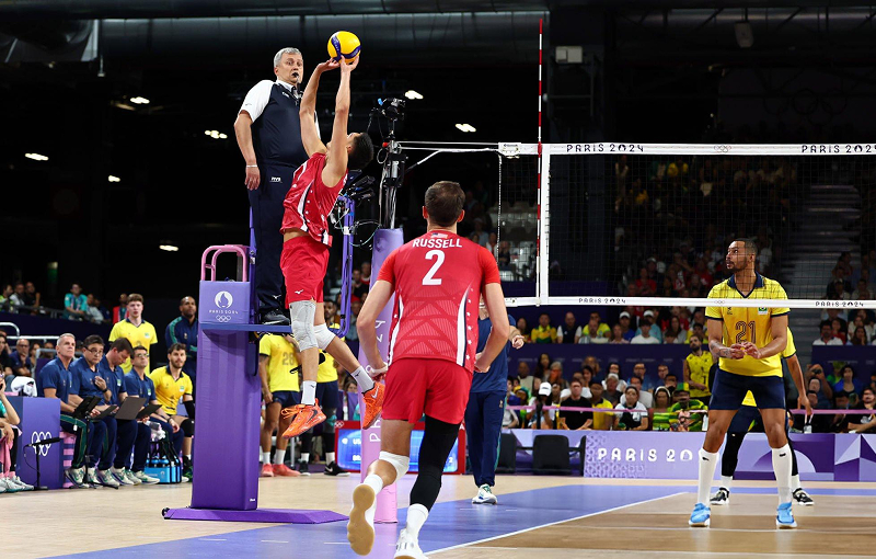 A volleyball player in a red uniform leaps to hit the ball over the net during a match. An official stands nearby, watching the play. Opponents in yellow prepare to defend. The court is marked with "Paris 2024."