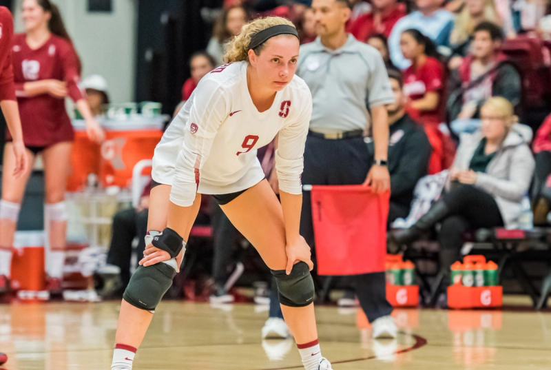 A volleyball player wearing a white jersey with the number 9 and knee pads is focused and leaning forward, ready for action on the court. A coach and audience are visible in the background.