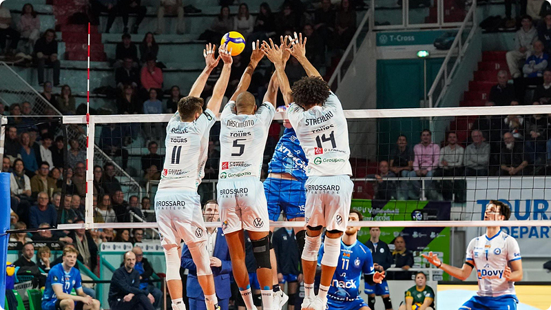 Players in white jerseys jump to block a volleyball at the net, as a player in a blue jersey attempts a spike. Spectators are visible in the background.