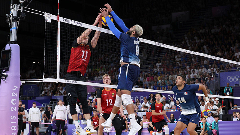 A volleyball match scene at the Paris 2024 Olympics shows a player in a blue jersey jumping to block the ball against an opponent in a red jersey at the net, with teammates and spectators in the background.