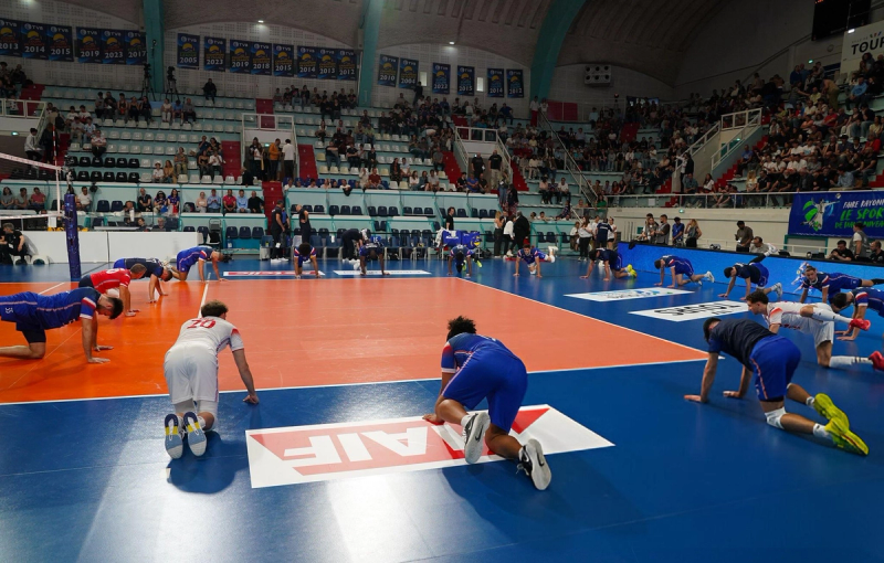 Volleyball players doing warm-up exercises on an indoor court with spectators in the stands.