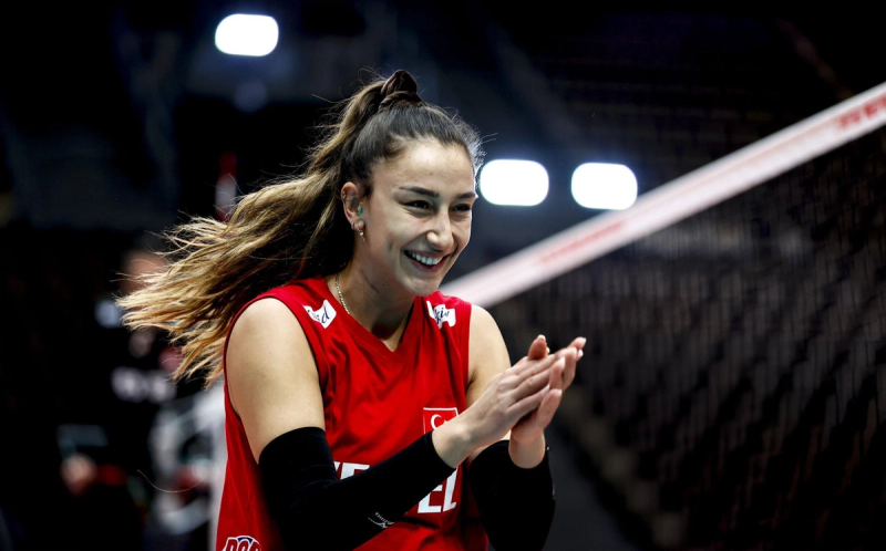 A smiling volleyball player in a red jersey clapping, with her hair tied back, under a sports hall light.