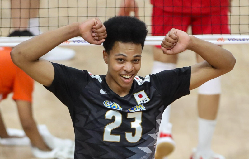 A volleyball player is smiling and flexing his arms in front of a net, wearing a black jersey with the number 23.
