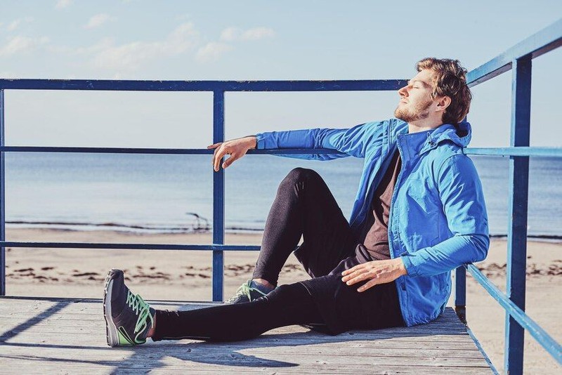A person in a blue jacket is sitting on a wooden deck by the beach, enjoying the sunlight with eyes closed. In the background, the sea and sand are visible.