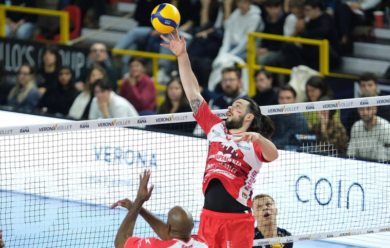 A volleyball player in a red and white uniform jumps to hit the ball over the net during a match. Spectators are seated in the background, watching intently.