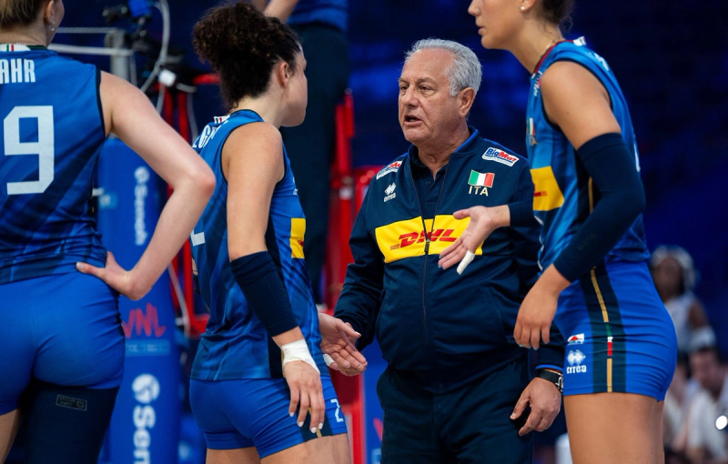 A volleyball coach speaks to players wearing blue uniforms with an Italian flag logo, during a match or timeout.