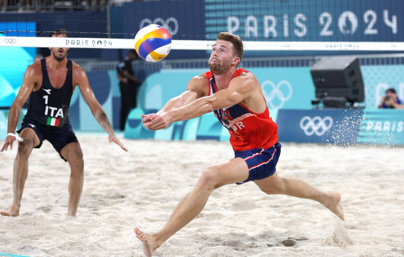 Two male beach volleyball players compete in the sand during the Paris 2024 Olympics. One player, wearing a red jersey, attempts a dig, while the other, in a dark jersey with "ITA" and an Italian flag, prepares to react. The Olympic rings are visible in the background.