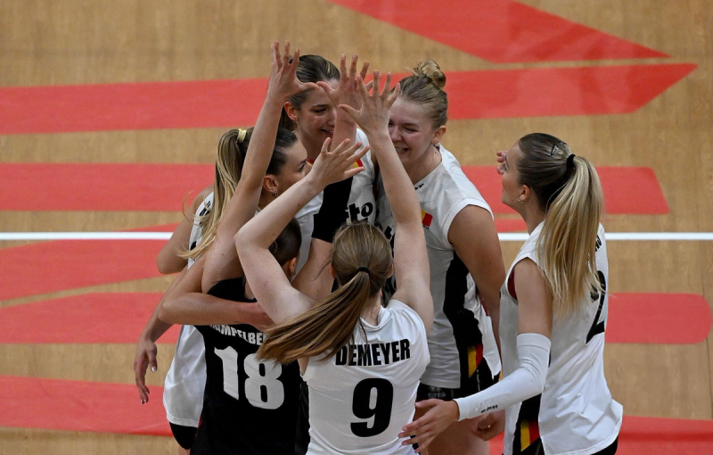 A group of female volleyball players celebrates on a court, raising their hands in a huddle and smiling.