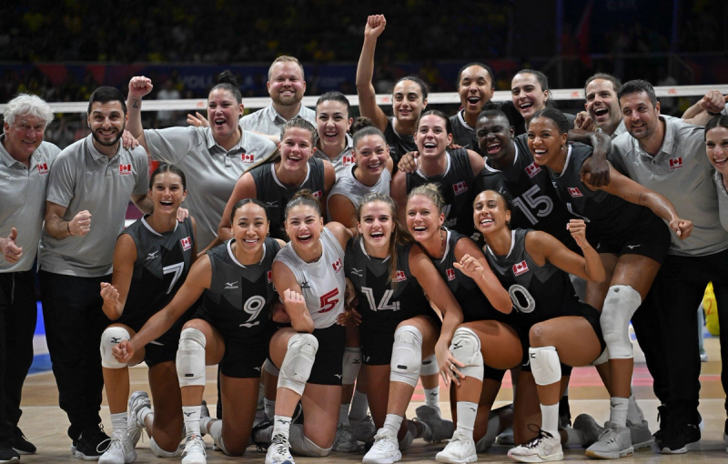 A jubilant group of female volleyball players and their coaches pose together on the court, wearing Canadian team jerseys. They are smiling and raising their fists in celebration.