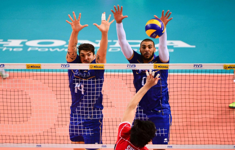 Two volleyball players in blue jerseys jump to block a ball over the net, while a player in a red jersey attempts to score.