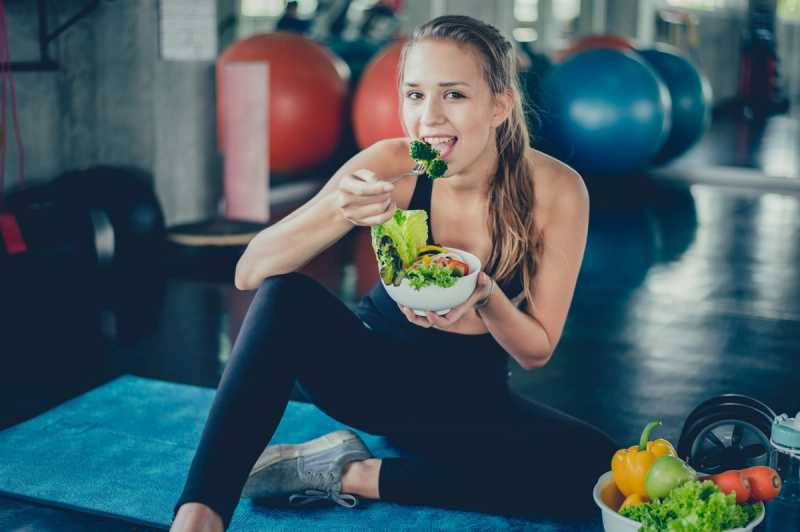 A person sitting on a blue mat in a gym, eating a salad with a fork. Exercise balls are visible in the background, and a bowl of fresh vegetables is nearby.