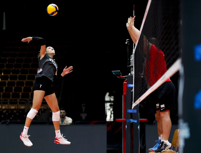 A volleyball player in a black uniform and numbered "7" jumps to spike the ball as an opponent in a red uniform attempts to block at the net.