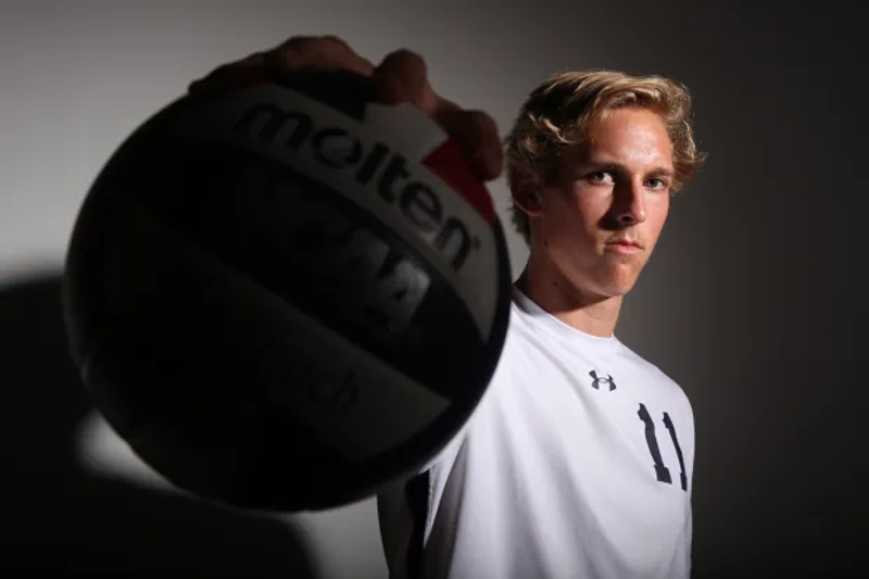 A person in a white jersey with the number 11 holds a volleyball towards the camera, casting a shadow on a plain background.