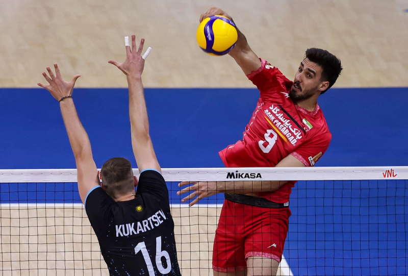 Two volleyball players in action near the net; one in a red uniform spiking the ball, and the other in a black uniform attempting a block.