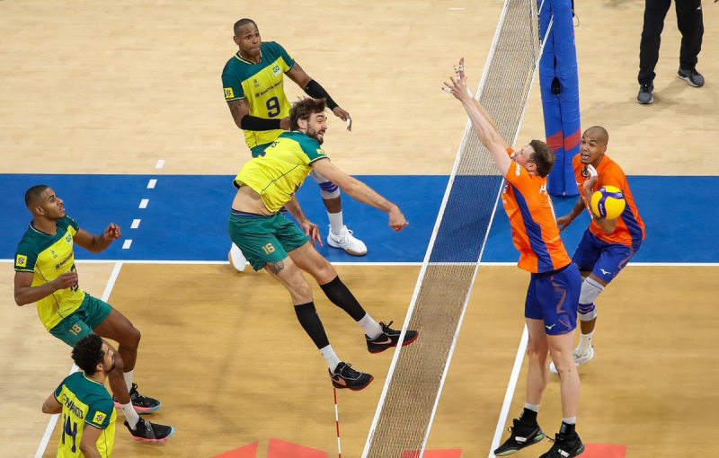 Two volleyball teams in action at the net. A player in a yellow and green jersey jumps to hit the ball while an opposing player in an orange jersey attempts to block. Other players observe the play.