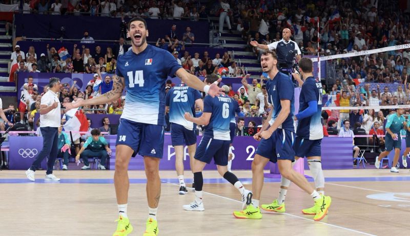 Volleyball players wearing blue and white celebrate energetically on the court, with a cheering crowd in the background during an Olympic event.