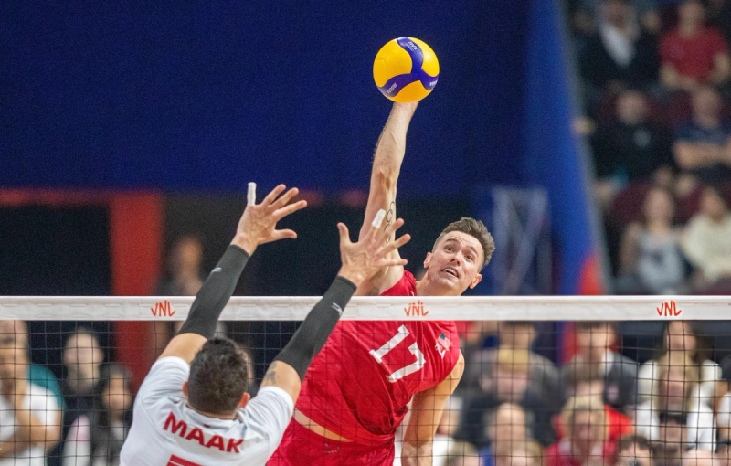 Two volleyball players at the net: one in a red jersey preparing to spike the ball, while the other in a white jersey attempts to block.