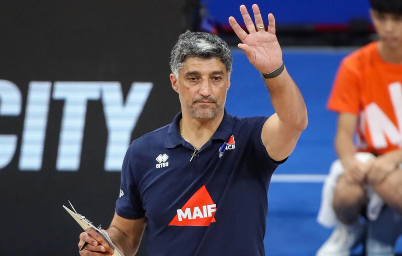 A man with gray hair wearing a navy sports polo is holding a clipboard and raising his hand. The background shows blurred spectators and a large screen displaying text.