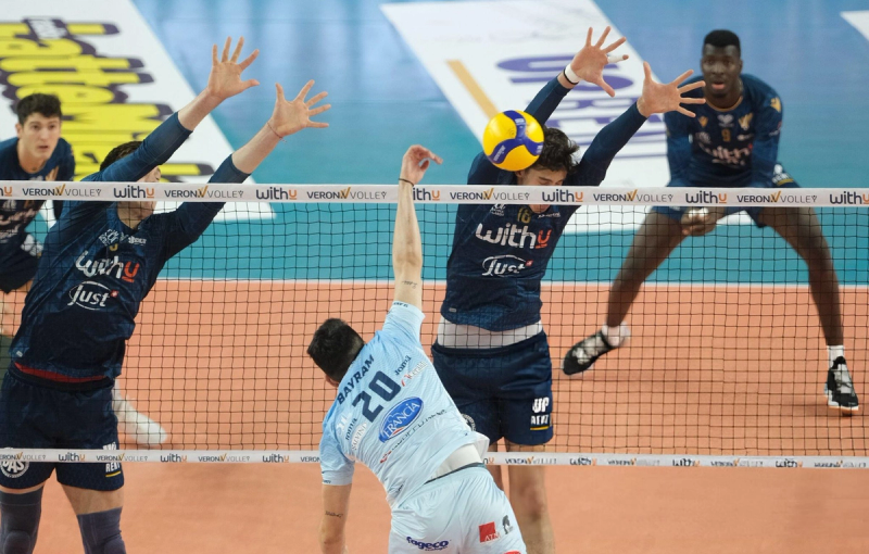 Volleyball players in action at the net, with two players in dark jerseys attempting to block a spike by a player in a light jersey, while another player prepares in the background.
