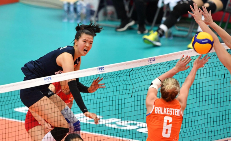 Two volleyball players from opposing teams are at the net. One, in a navy uniform, is spiking the ball, while the other, in an orange uniform, attempts to block it.