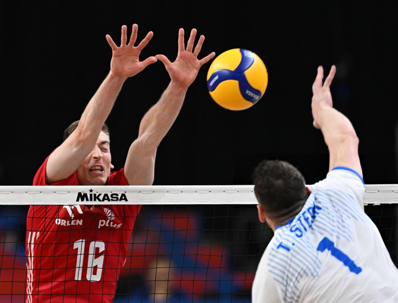 A volleyball player in a red jersey attempts to block a spike from an opposing player in a white jersey. The ball is seen above the net.