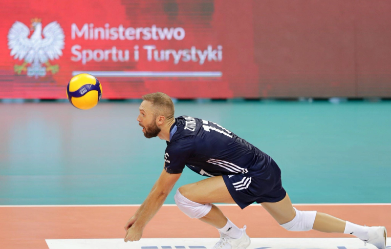 A volleyball player in a navy uniform lunges forward to hit a ball. The background features a red banner with the logo of the Ministry of Sport and Tourism.