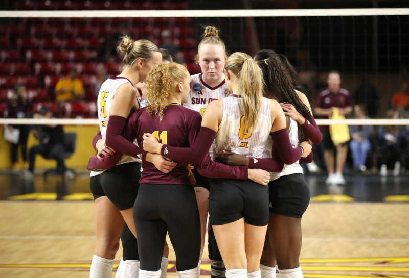 A volleyball team huddles on the court with arms around each other, wearing matching uniforms.