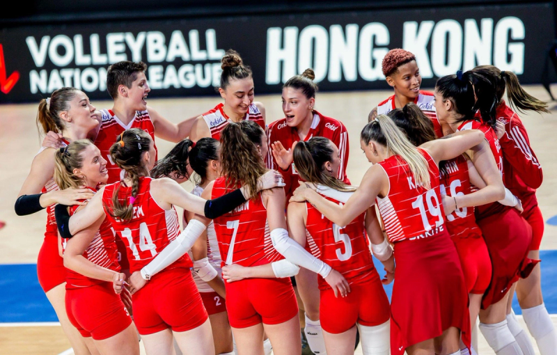 A volleyball team, wearing red uniforms, huddles together on the court, smiling and cheering. A "Volleyball Nations League" sign is visible in the background.