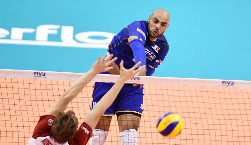 A volleyball player in a blue uniform jumps and spikes the ball over the net, while another player in a red uniform attempts to block.