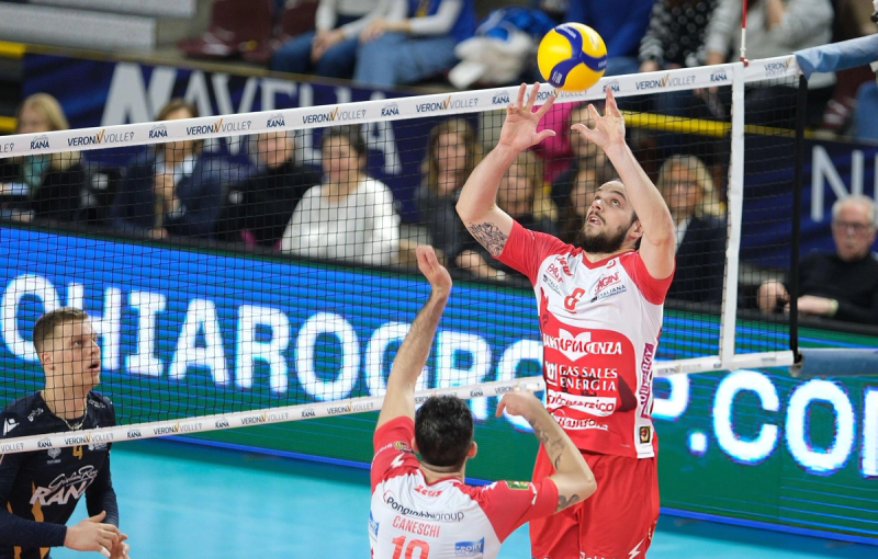 Volleyball player in a red and white uniform sets the ball during a match, with another player in a dark uniform looking on. The net displays "VERONA VOLLEY." Spectators are visible in the background.