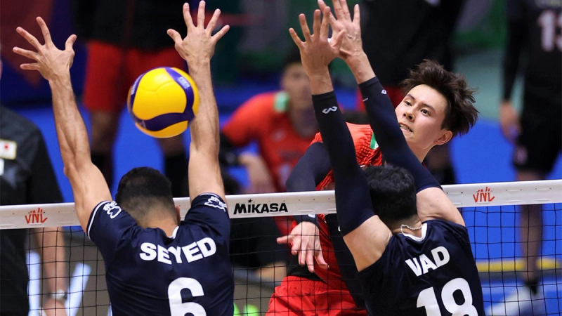 Two volleyball players in navy jerseys attempt to block a spike from a player in a red jersey during a match. The ball is seen in mid-air above the net.