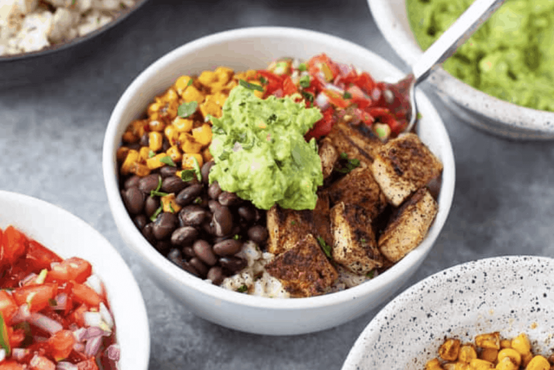 A bowl filled with seasoned tofu, black beans, grilled corn, guacamole, and tomato salsa, surrounded by other bowls of toppings.