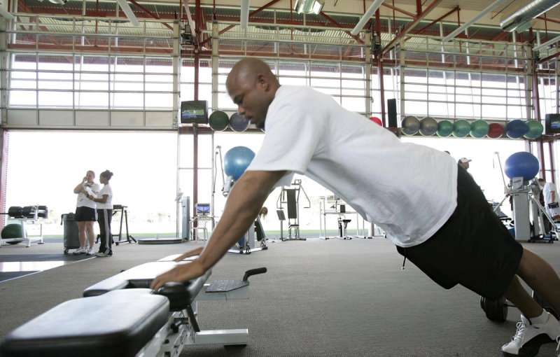 A person in a white t-shirt and black shorts is doing inclined push-ups using a bench in a gym. The gym has large windows, exercise equipment, and several medicine balls visible in the background. Two other people are standing and talking in the distance.