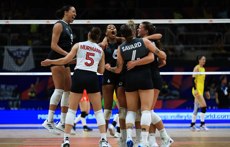 A women's volleyball team celebrates energetically on the court, with players in black uniforms embracing and smiling. One player in a white uniform joins in while an opposing team member in a yellow uniform walks by in the background.