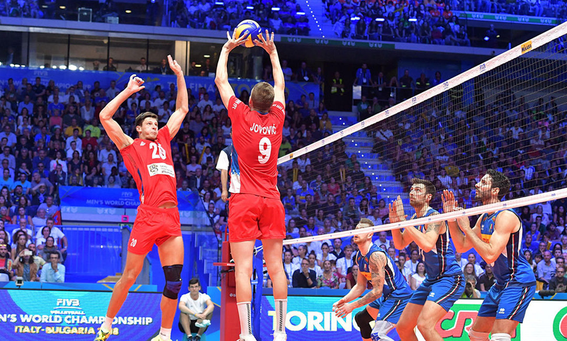 A men's volleyball match scene where players in red are engaged in setting the ball while players in blue anticipate on the other side of the net. The match takes place in a large indoor arena with a crowd watching.