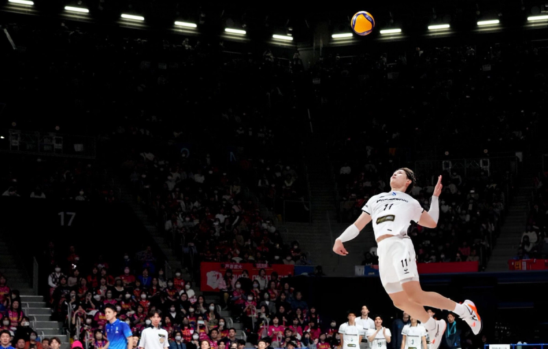 A volleyball player in a white uniform leaps into the air, preparing to serve the ball in an indoor stadium filled with spectators.