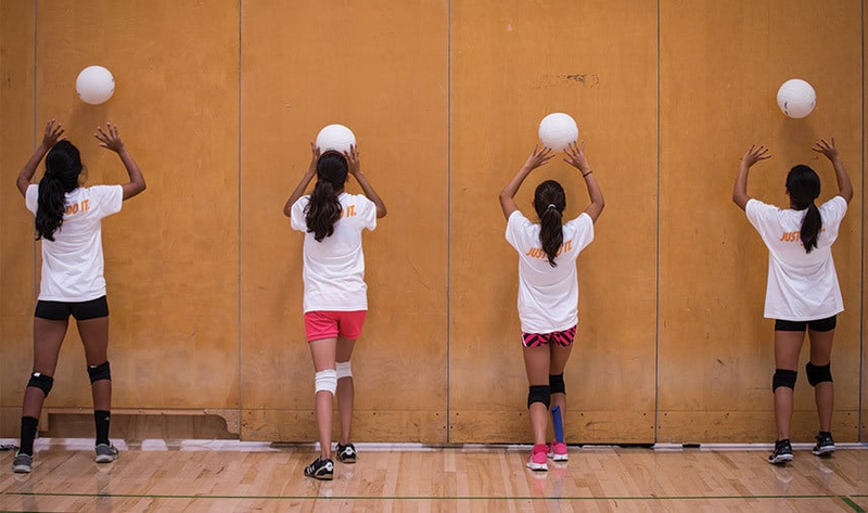 Four girls in athletic outfits practice volleyball drills by holding balls against a gym wall.