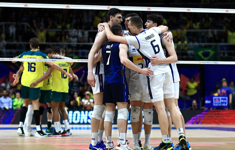 Volleyball players in white uniforms huddle together on the court. In the background, players in yellow uniforms also huddle. A volleyball net separates the teams, and the crowd is visible in the stands.