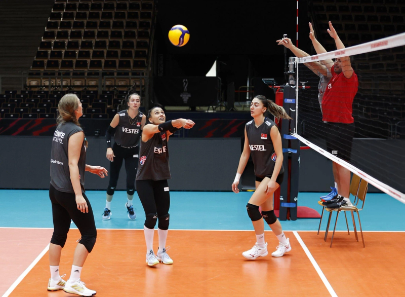 A women's volleyball team practices on the court, with one player performing a bump while others watch. A coach stands by the net.