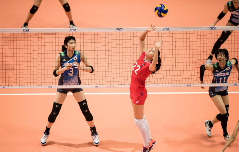 A volleyball player in a red uniform sets the ball near the net, while three opposing players in blue uniforms prepare to react on a court.