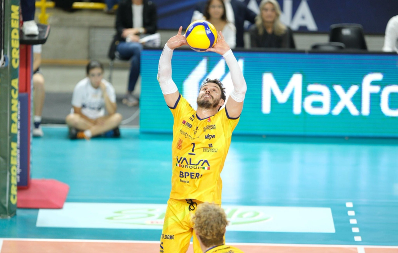 A volleyball player in a yellow uniform sets the ball during a match. The court is blue and there are spectators in the background.