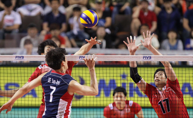 Two volleyball players are in action at the net, with one player jumping to spike the ball and the opposing player attempting to block. The scene takes place during a competitive match, with spectators visible in the background.