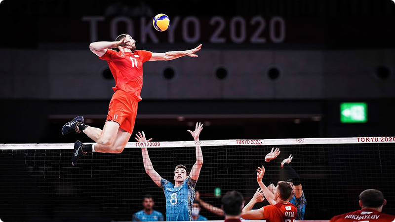 A volleyball player in a red uniform jumps high to spike the ball over the net, while two opposing players in blue jerseys attempt to block. The setting is the Tokyo 2020 Olympic Games.