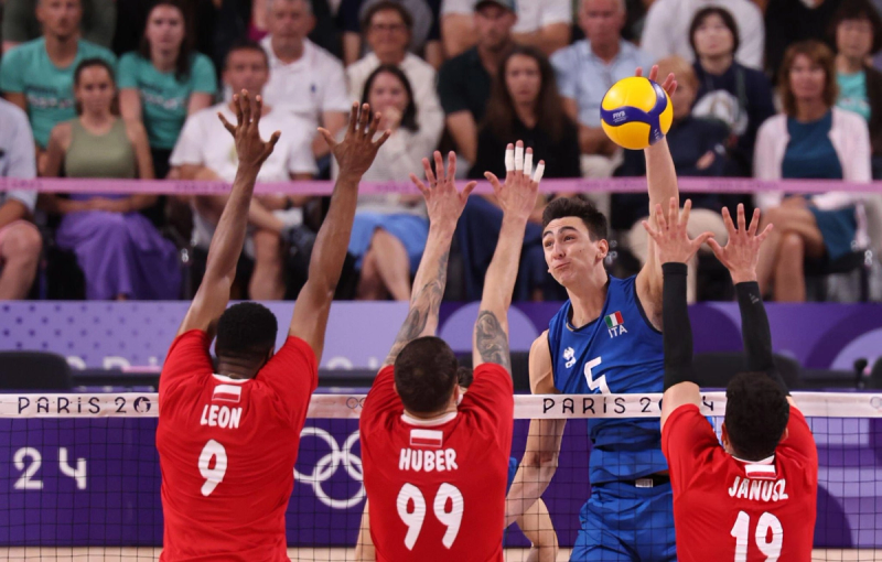 A volleyball player in a blue jersey is spiking the ball over the net, while three opposing players in red jerseys attempt to block. Spectators are visible in the background.