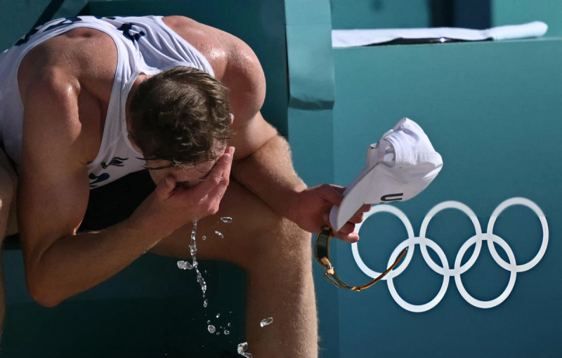 An athlete in a white uniform sits, holding a cap and wiping sweat from their face. The Olympic rings are visible in the background.