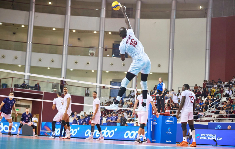 A volleyball player in a white uniform jumps high to spike the ball during a match in an indoor arena, with teammates and opponents watching.