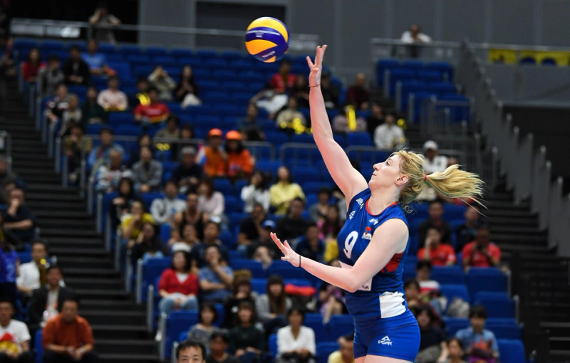 Volleyball player wearing a blue and white uniform, number 9, poised to serve the ball in a stadium with spectators in the background.