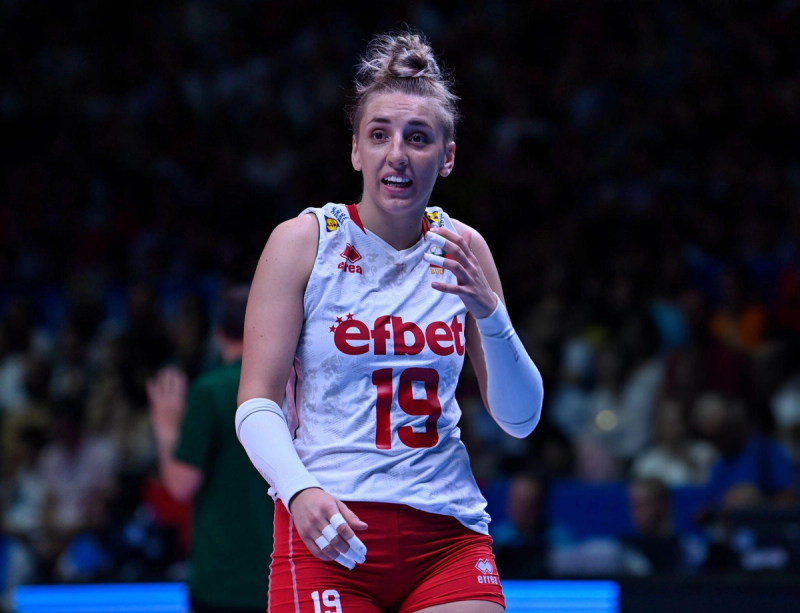 Volleyball player wearing a white jersey with the number 19 and the word "efbet," gesturing during a game, with a blurred audience in the background.
