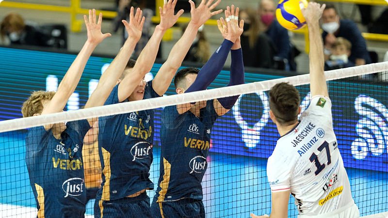 A volleyball player in a white jersey prepares to spike the ball over the net while three players in blue jerseys attempt to block it.