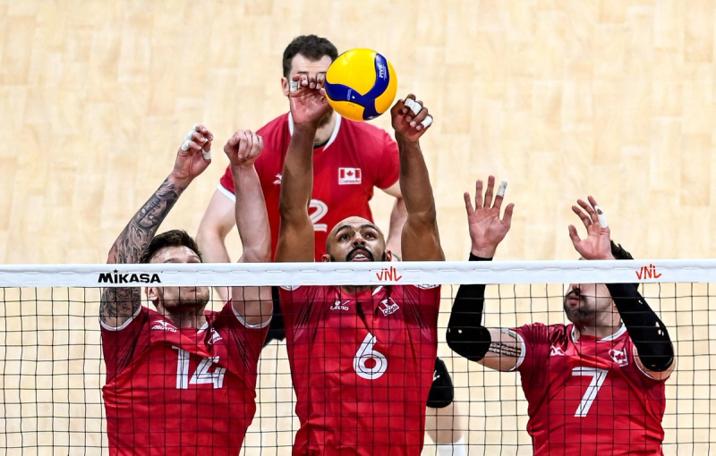 Three volleyball players in red jerseys jump near the net to hit a ball.